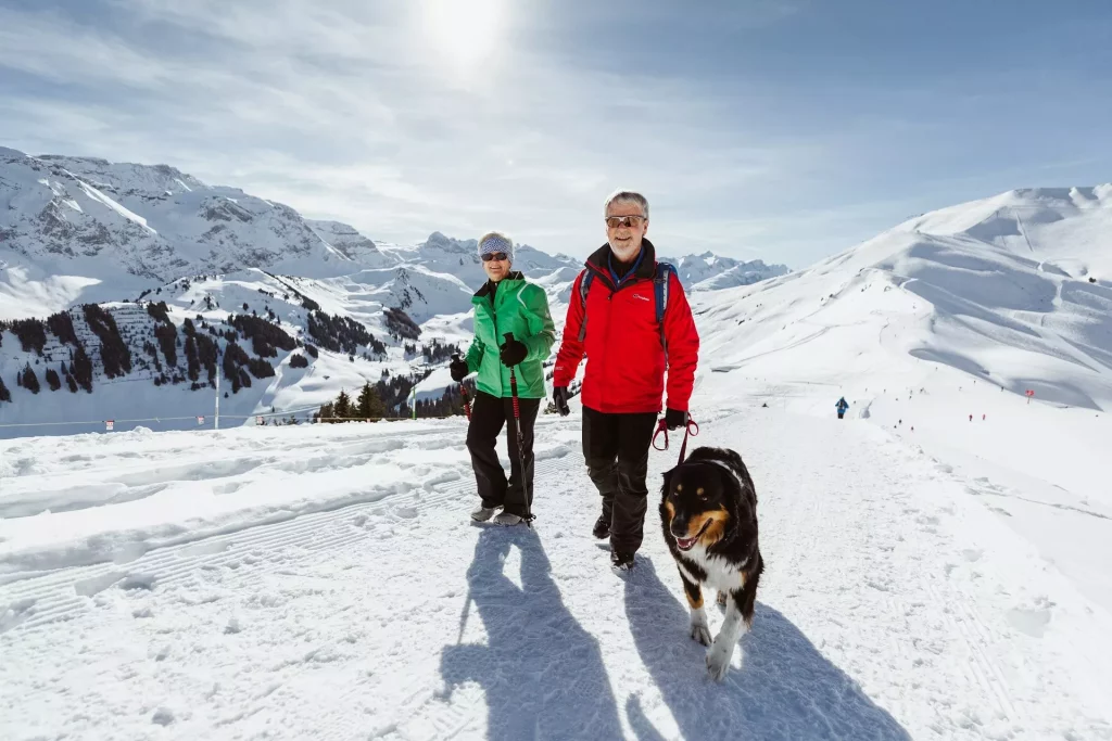 Älteres Paar wandert mit Hund auf verschneitem Weg in den Bergen bei Sonnenschein